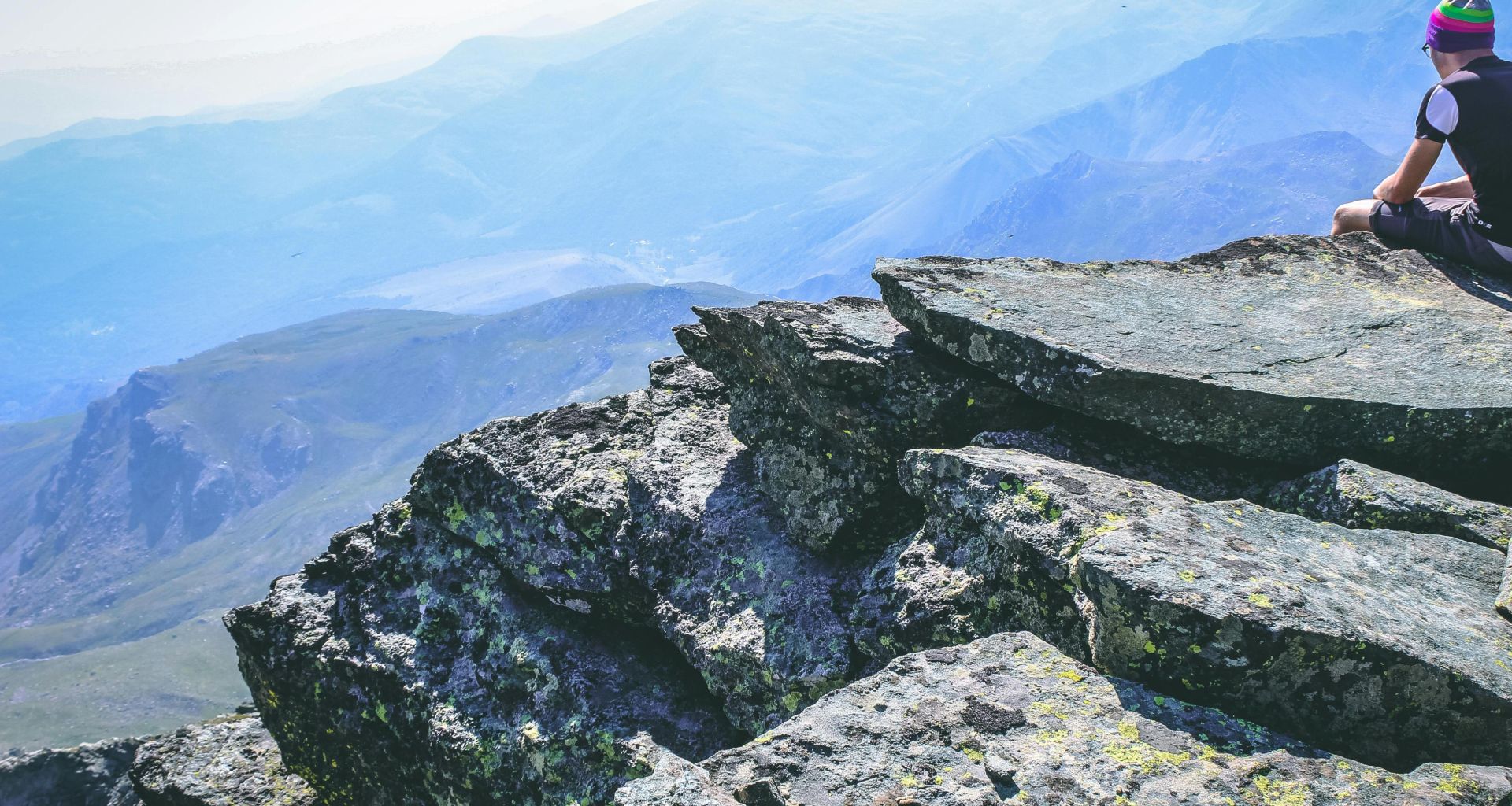 A lone hiker in colorful hat sits on a rocky mountain peak, enjoying the scenic view.