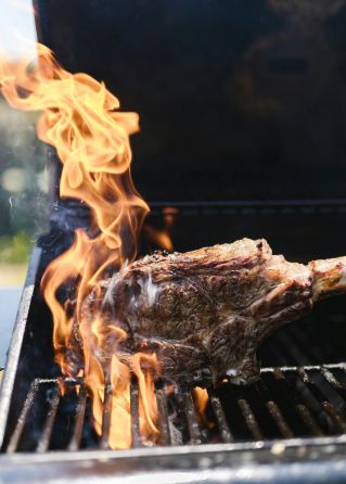 A sizzling tomahawk steak on a barbecue grill with vibrant flames during a sunny outdoor cookout.